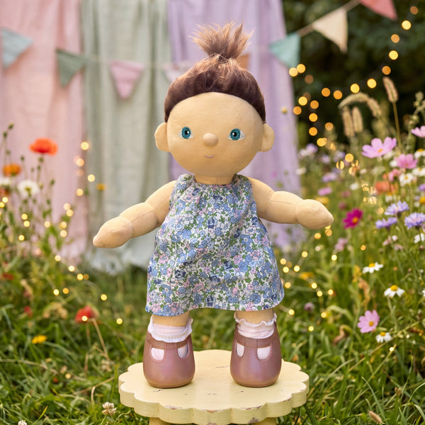 Doll in a floral dress standing on a wooden platform in a garden setting with flowers and lights.