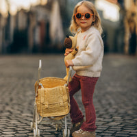 Child in orange sunglasses holding a teddy bear beside a honey yellow doll stroller with an attached changing bag from Dinkum Dolls