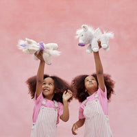 Two smiling children in pink shirts and striped overalls hold plush unicorn toys overhead against a pink backdrop, featuring the sparkly Dinkum Unicorn Glimmer