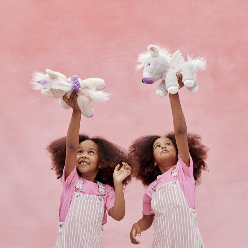 Two smiling children in pink shirts and striped overalls hold plush unicorn toys overhead against a pink backdrop, featuring the sparkly Dinkum Unicorn Glimmer