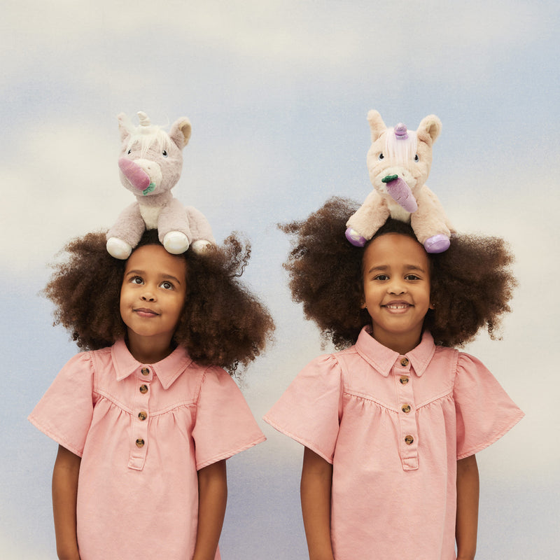 Two smiling children in pink dresses pose with soft lilac unicorn toys on their heads from Dinkum Unicorns in a playful studio scene