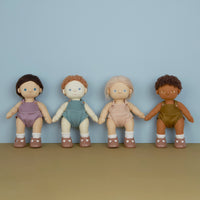 Four Dinkum Dolls with diverse skin tones and hairstyles stand side by side in colorful bodysuits against a soft blue wall.