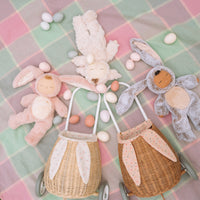 Two rattan bunny luggys with floppy ears and pastel speckled linings, surrounded by plush bunnies and colorful Easter eggs on a soft plaid blanket by Olli Ella Kids Rattan