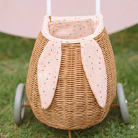 Rattan bunny luggy basket with speckled Gumdrop lining and floppy fabric ears on wheels, shown outdoors on grass by Olli Ella Kids Rattan