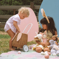 Two children play outdoors beside a rattan bunny luggy with white wheels and speckled pink lining, surrounded by plush toys and pastel balls.