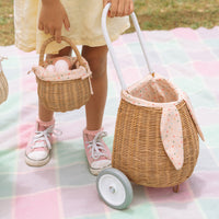Child holding a small rattan basket beside an Olli Ella Kids Rattan bunny luggy with speckled Gumdrop lining and long bunny ears on a pastel blanket