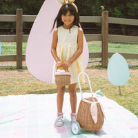 Young girl holding a small rattan basket beside an Olli Ella bunny luggy with wheels, floppy ears, and a pink speckled lining outdoors on a picnic blanket