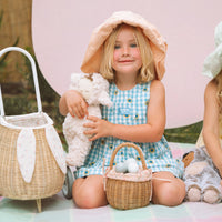 Young girl holding a plush bunny beside an Olli Ella Kids Rattan bunny luggy with floral lining and a small woven basket in front