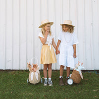 Two children in sun hats stand outdoors beside rattan bunny luggy baskets, including a floral-lined Pansy bunny luggy by Olli Ella Kids Rattan.