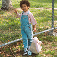 Child in denim overalls holding an Olli Ella Kids Rattan Bunny Luggy with pink floppy ears and wheels, shown outdoors beside a chain link fence.
