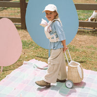 Child in a sunhat pulls an Olli Ella Kids Rattan bunny luggy with floral Pansy lining across a pastel picnic blanket outdoors.