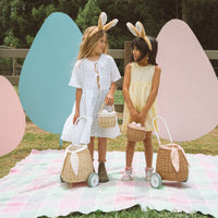 Two children wearing bunny ear headbands pose outdoors with wheeled rattan bunny baskets including a Pansy lined basket and a natural brown basket by Olli Ella Kids Rattan