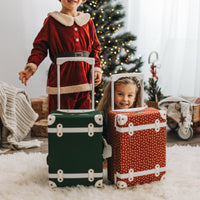 Two children with green and red rolling suitcases beside a Christmas tree, featuring the Forest See-Ya Suitcase by Olli Ella Kids for festive travel and sleepovers
