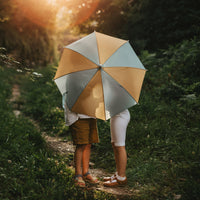 Two children walk along a sunlit woodland path beneath a blue and butter yellow See Ya umbrella by Olli Ella Kids
