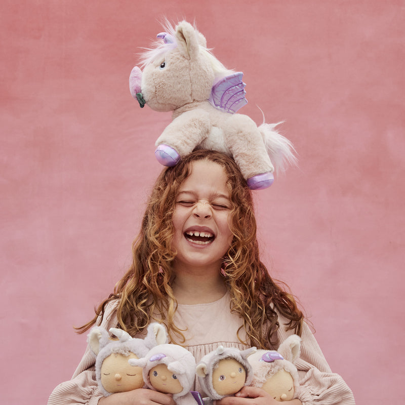 Child laughing while wearing a plush pink unicorn on her head and holding three soft dolls, showcasing the playful Dinkum Pets Unicorn Shimmer toy collection