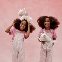 Two smiling girls in pink striped overalls play with plush pink and white unicorn toys from Dinkum Pets against a soft pink background
