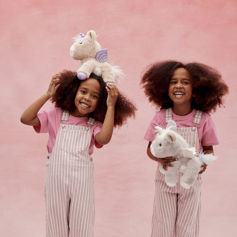 Two smiling girls in pink striped overalls play with plush pink and white unicorn toys from Dinkum Pets against a soft pink background