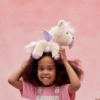 Smiling child holding a soft pink Dinkum Unicorn Shimmer plush on her head, showing its lavender wings, fluffy mane, tail, and purple hooves against a pink backdrop