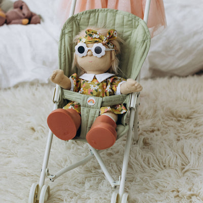 Dinkum Dolls doll wearing a floral outfit seated in a sage green stroller seat with a safety bar and white frame indoors on a soft rug