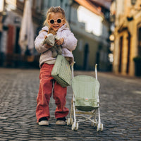 Young child in sunglasses beside a sage green doll stroller seat and matching bag on a cobbled street, showcasing Dinkum Dolls stroller accessories