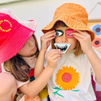 Two children play with Olli Ella Kids Tubbles Funspressions, holding interchangeable eye, nose, and smiling mouth pieces to create funny faces.
