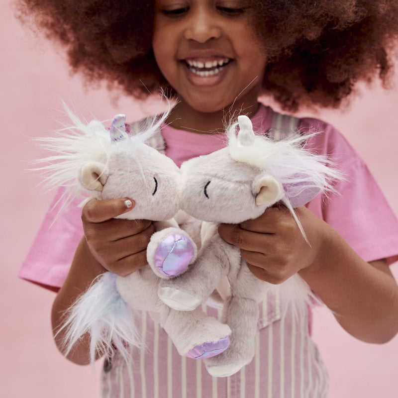 Smiling child holding two soft white unicorn plush toys with fluffy pastel manes and glittery lilac details from Dinkum Pets
