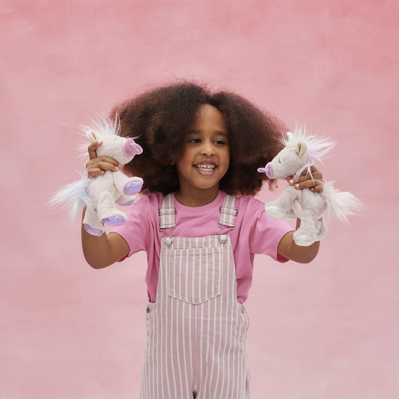 Smiling child in pink clothing holding two soft unicorn toys, including a lilac glitter unicorn from Dinkum Pets, against a pink backdrop