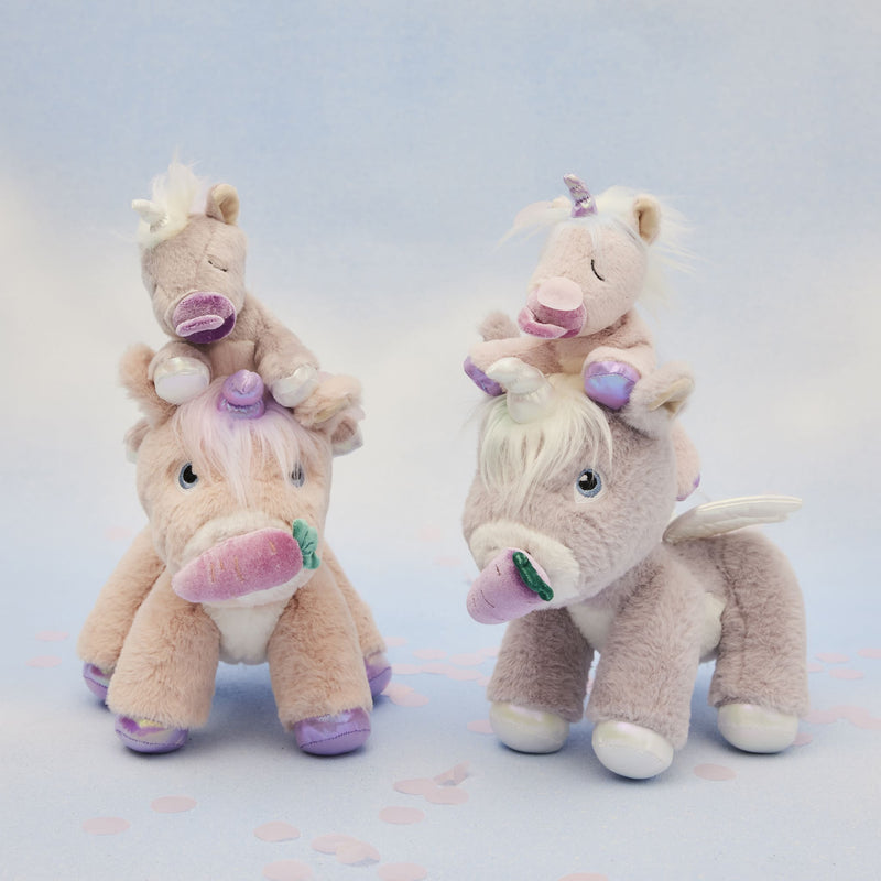 Two pastel plush unicorn toys, one pink and one lilac, each carrying a tiny unicorn on its back, with soft fur, colorful horns, wings, and embroidered eyes from Dinkum Pets