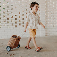 Young child pulling a natural rattan wheeled basket along a paved walkway, showcasing the Olli Ella Kids Rattan Original Luggy for play and storage.