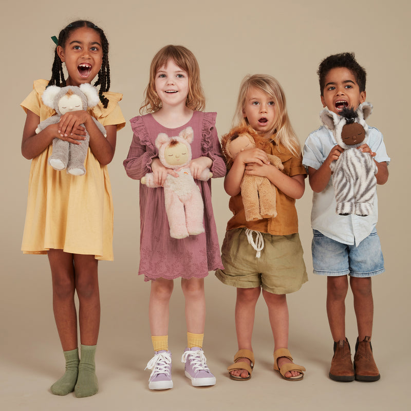 Four smiling children pose together while holding Cozy Dinkums plush toys, including a zebra in a soft striped suit, in a warm studio setting.