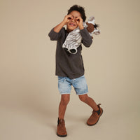 Child playfully wearing a Cozy Dinkums Zebra Mini plush toy around their neck while posing in a dark shirt, denim shorts, and brown boots