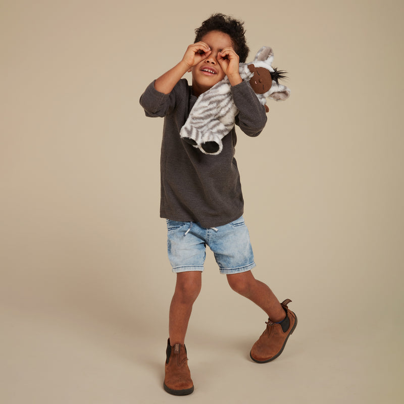 Child playfully wearing a Cozy Dinkums Zebra Mini plush toy around their neck while posing in a dark shirt, denim shorts, and brown boots