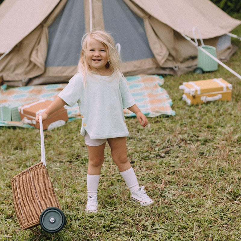 A smiling child pulls a natural rattan wheeled Luggy basket across grass at a campsite, with a tent and picnic blanket behind, by Olli Ella Kids Rattan