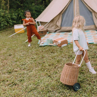 Child pulling a natural rattan wheeled basket across the grass at a campsite, with a tent and another child playing in the background