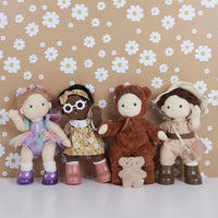 Four Dinkum Dolls pose together in colorful outfits, including a fairy doll in a purple tutu with wings, alongside a small teddy bear against a floral wall.