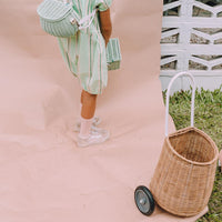 Natural rattan wheeled Luggy basket beside a child in pastel clothing, with a tall handle and sturdy wheels for carrying toys, treasures, or picnic essentials.