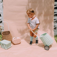 Toddler pulling a pale green wheeled rattan luggy basket outdoors, with natural woven baskets nearby from Olli Ella Kids Rattan