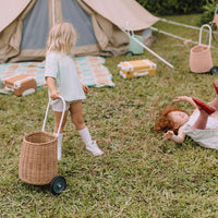 Child pulling a natural rattan wheeled basket across grass at a campsite, with a tent and picnic toys in the background from Olli Ella Kids Rattan
