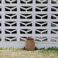 Small natural rattan wheeled basket with a tall handle resting on grass against a white breeze block wall, by Olli Ella Kids Rattan
